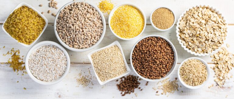 Selection of whole grains in white bowls on white wood background.