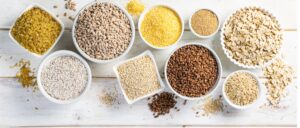 Selection of whole grains in white bowls on white wood background.